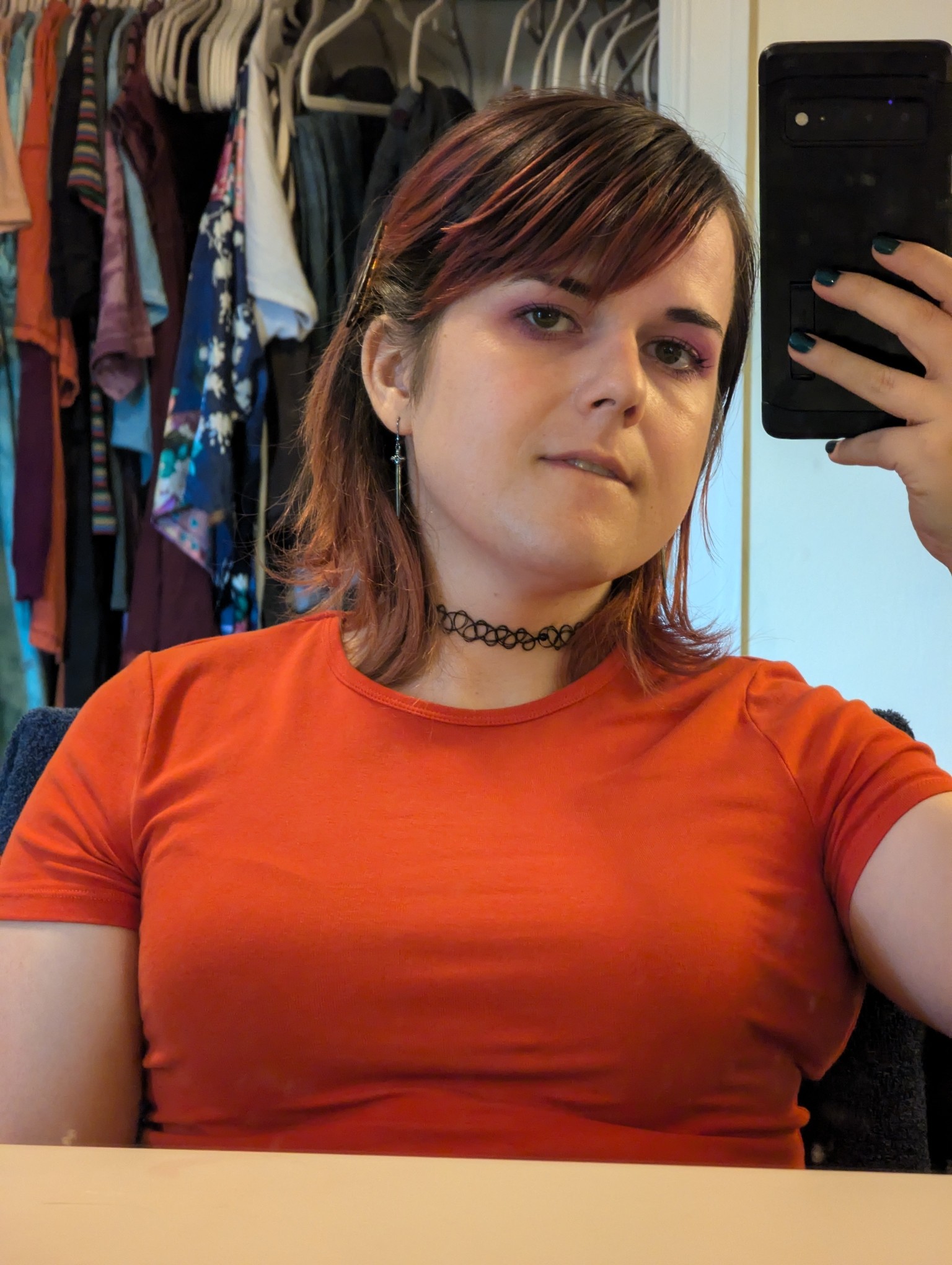 a girl in an orange top, with red hair, and a black choker. she's looking into the camera while seated.