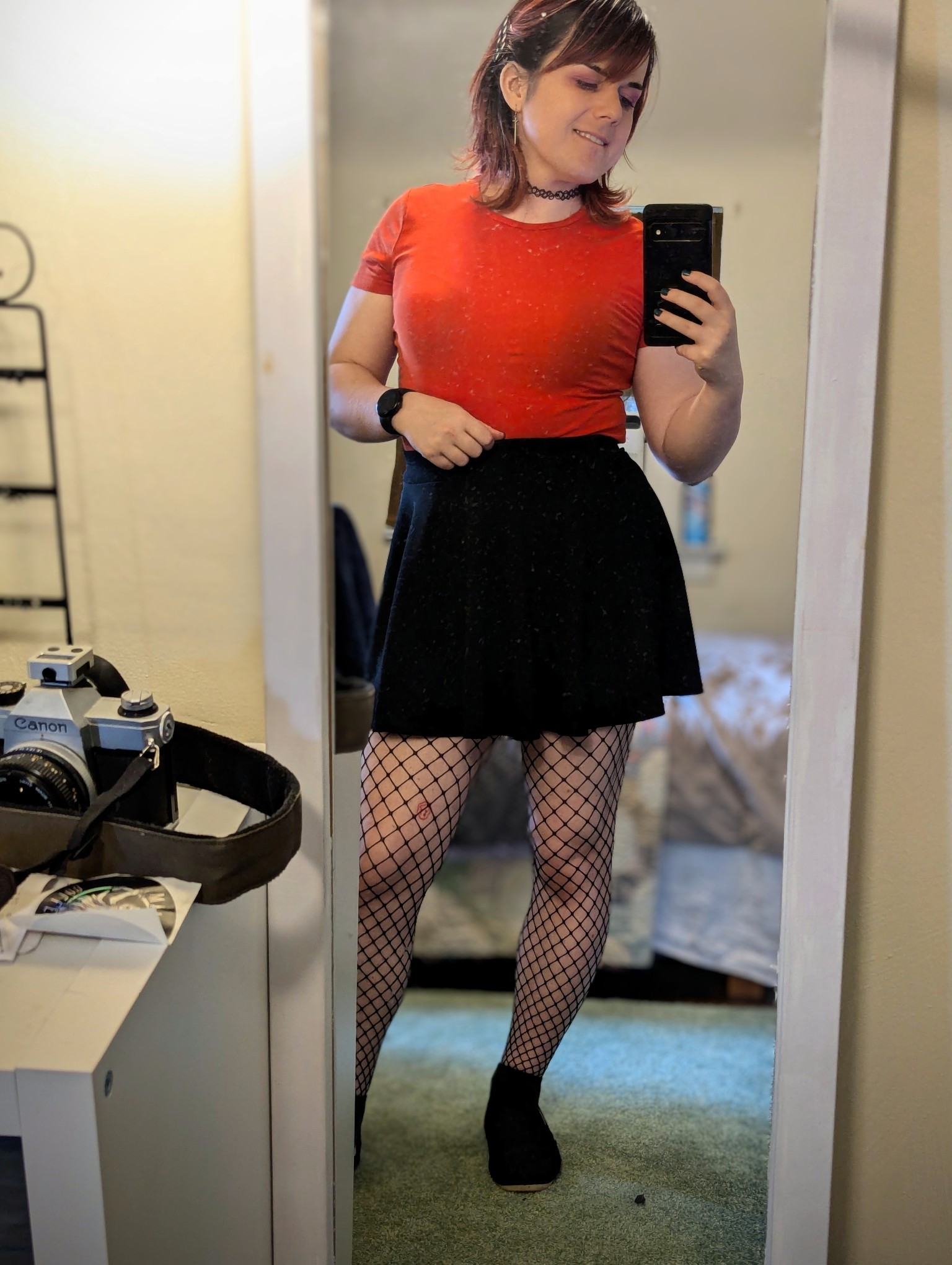 A girl taking a picture in a mirror. She's wearing an orange fitted top, a black skirt, a black choker, and fishnets. She's smiling and looking at her phone.