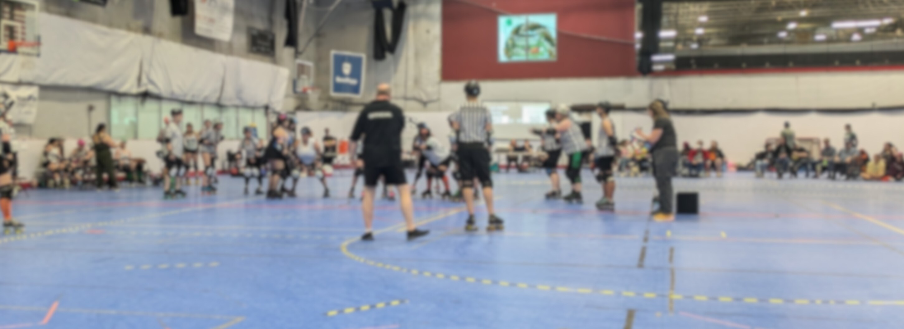 A blurred image of a roller Derby bout inside a large indoor gymnasium. A group of people are lined up and two groups of referees stand by, ready to begin the jam. Several people sit on the sidelines in folding chairs.