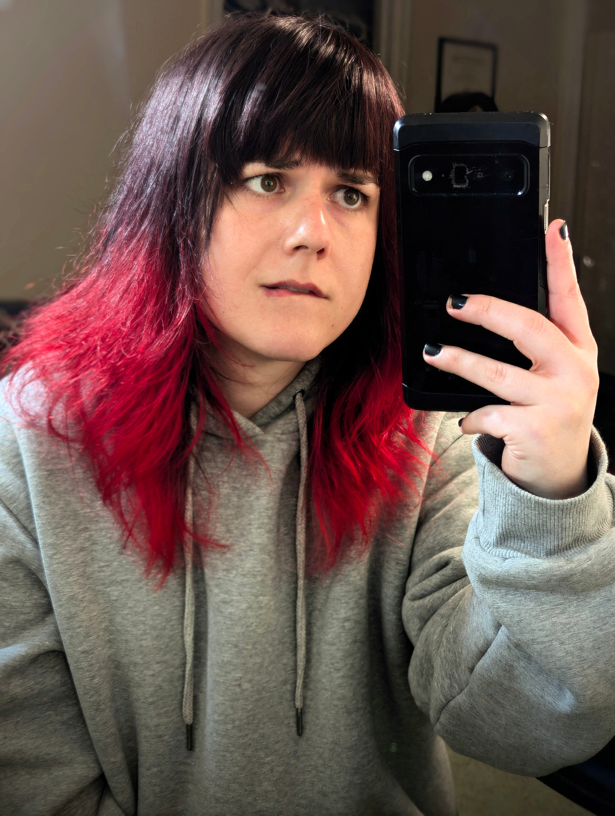 A girl with collarbone-length hair with a natural dark brown to vibrant red ombre. She's staring at her phone while biting her lip, and making a slightly concerned looking expression. She's wearing a grey oversized pullover.