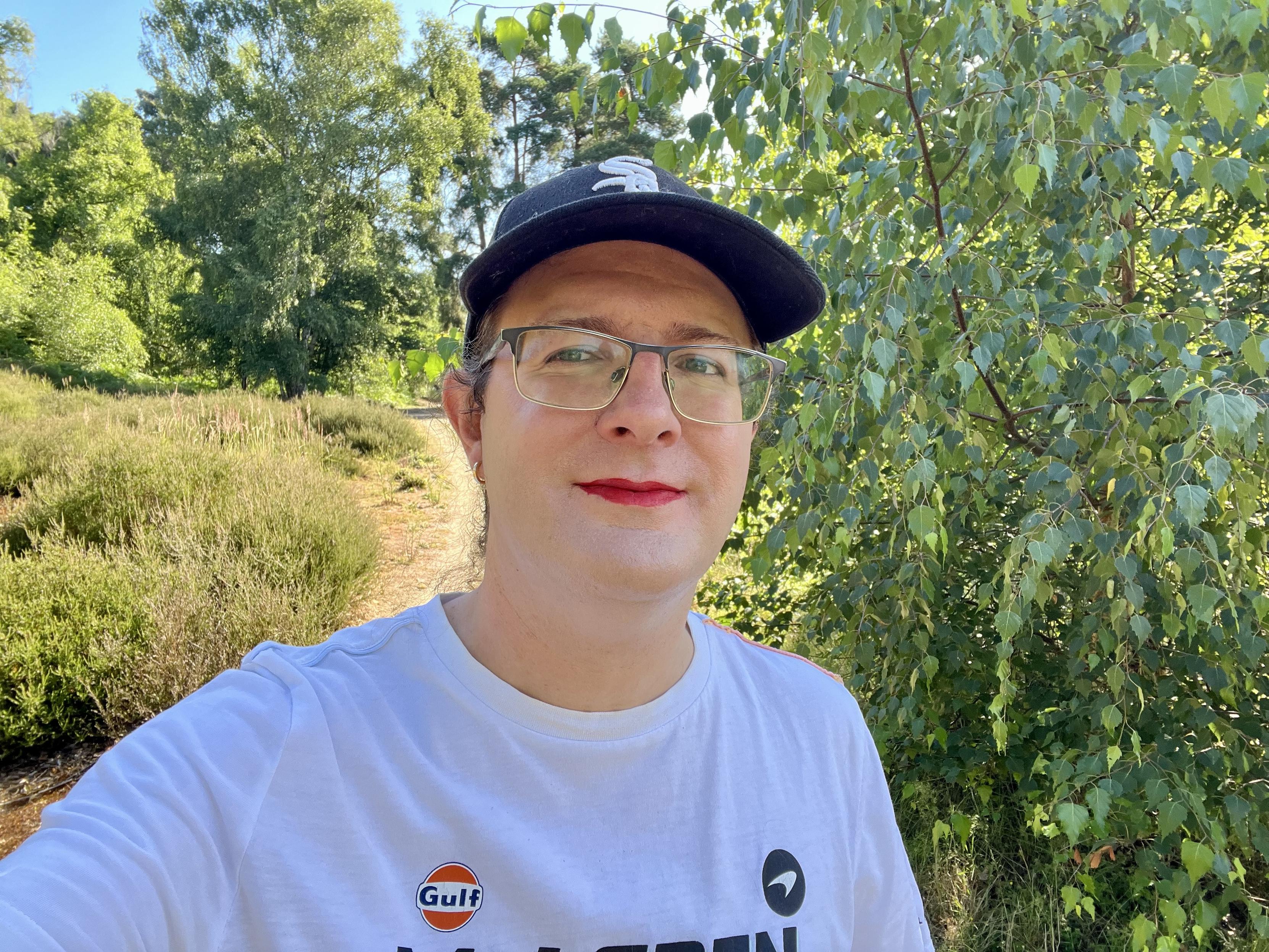 A selfie of Ellie, who is looking at the camera while standing on a sandy path next to some bushes on a bright, sunny day. 

She’s wearing a black Chicago White Sox baseball cap, glasses, small gold hoop earrings, and a white McLaren Racing t-shirt.