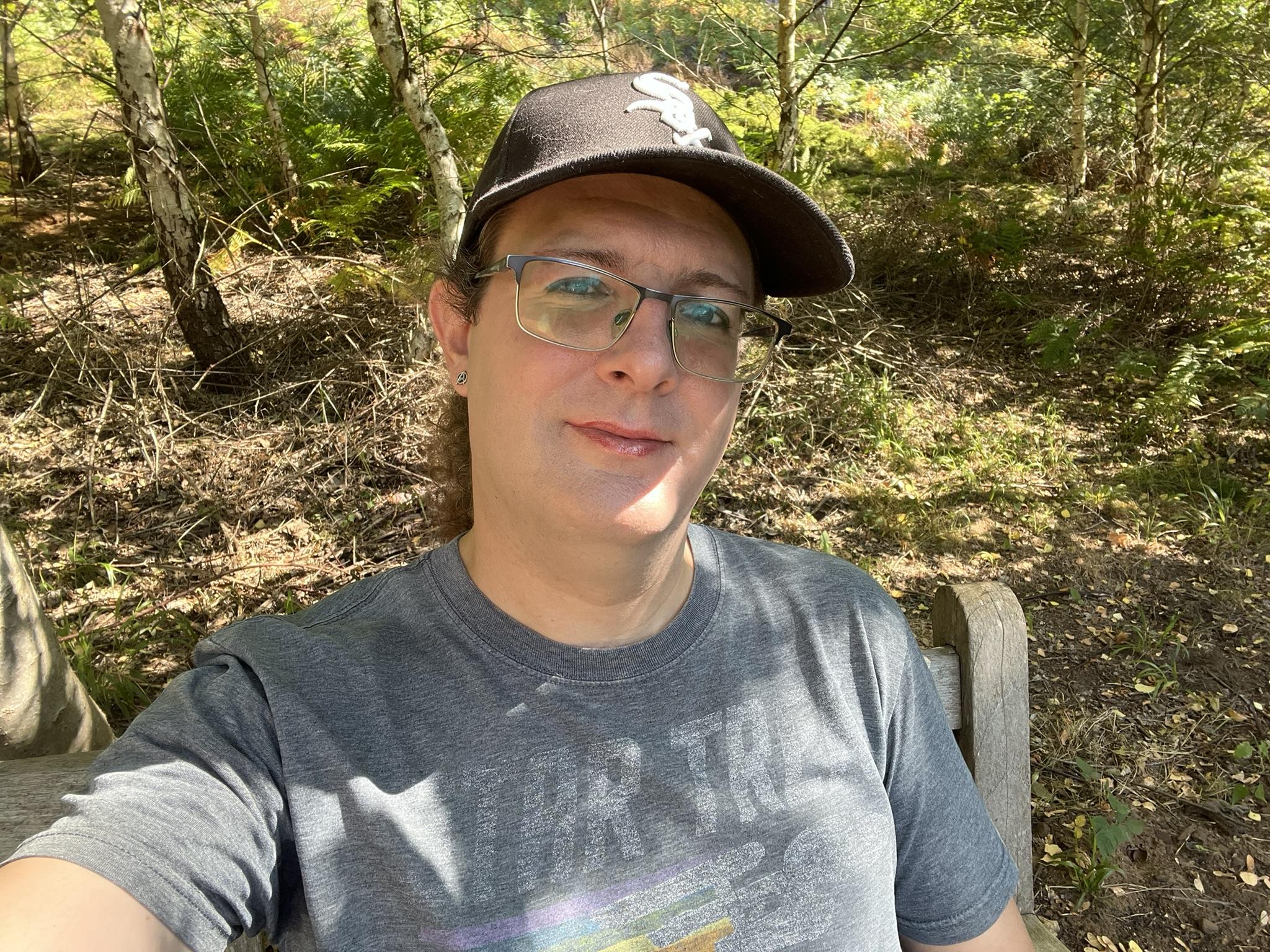 A selfie of Ellie, who's sitting on a wooden bench in front of some trees and greenery.

She's wearing a Chicago White Sox baseball cap (on a 3-win run, including two against the Yankees, just so you know), glasses, Starfleet delta earrings, and a grey t-shirt with a faded retro-style Star Trek print, with the original series logo at the top and a silhouette of the Enterprise leaving a rainbow trail behind it.